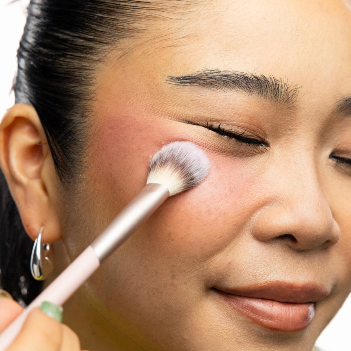 Lähikuva henkilöstä, joka levittää poskipunaa pyöreällä meikkisiveltimellä poskelle. Henkilöllä on tumma hiusnuttura ja hopeanväriset korvakorut.