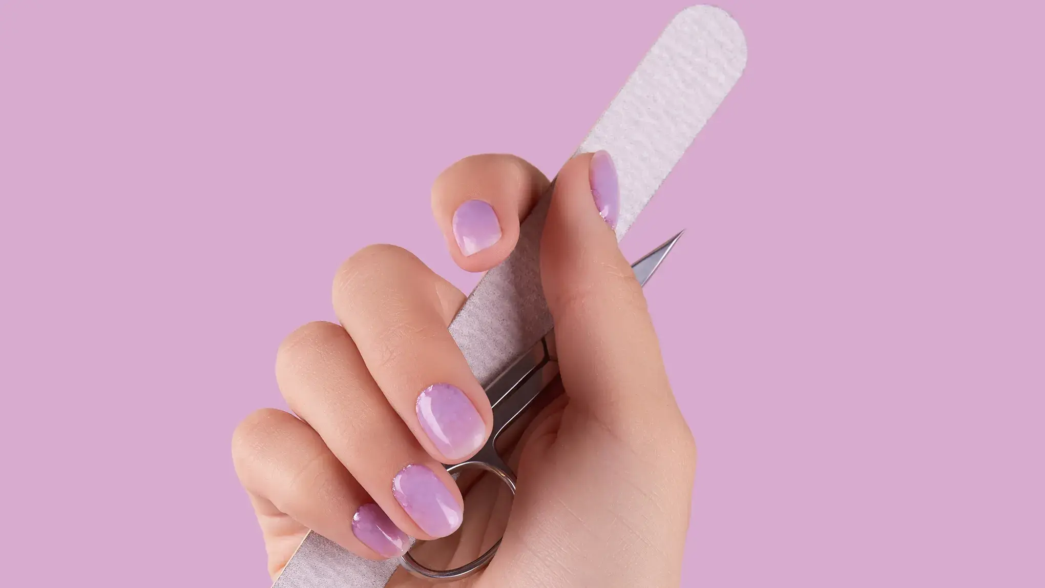 Hand som håller en nagefil och en nagelsax. Naglarna är lila och bakrgunden lila.
