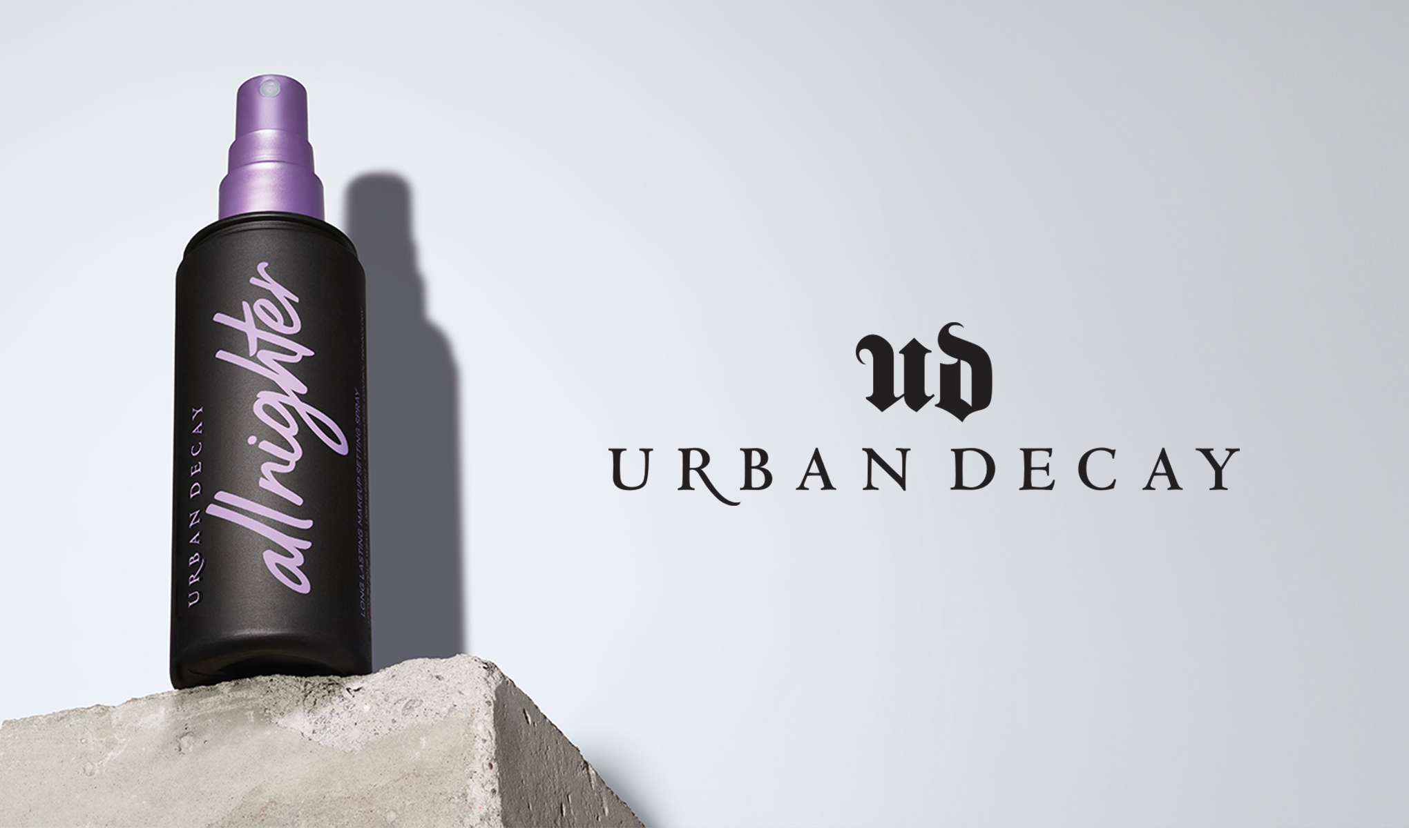 En flaska Urban Decay All Nighter Setting Spray med lila spraymunstycke står på en betongblock mot en vit bakgrund. Till höger i bilden syns texten "UD Urban Decay".