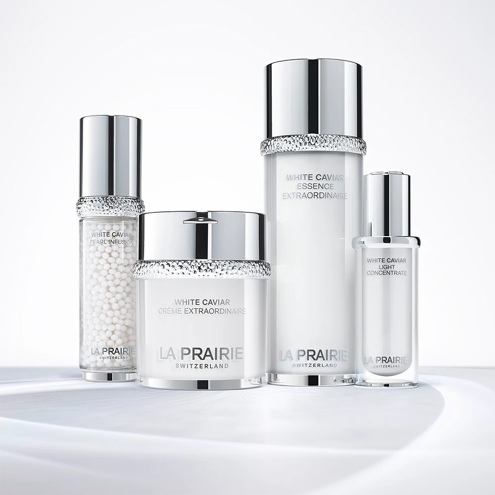 La Prairie White Caviar -ihonhoitotuotteet hopeanvalkoisissa pulloissa ja purkissa, aseteltuna kirkkaalle ja vaalealle alustalle.