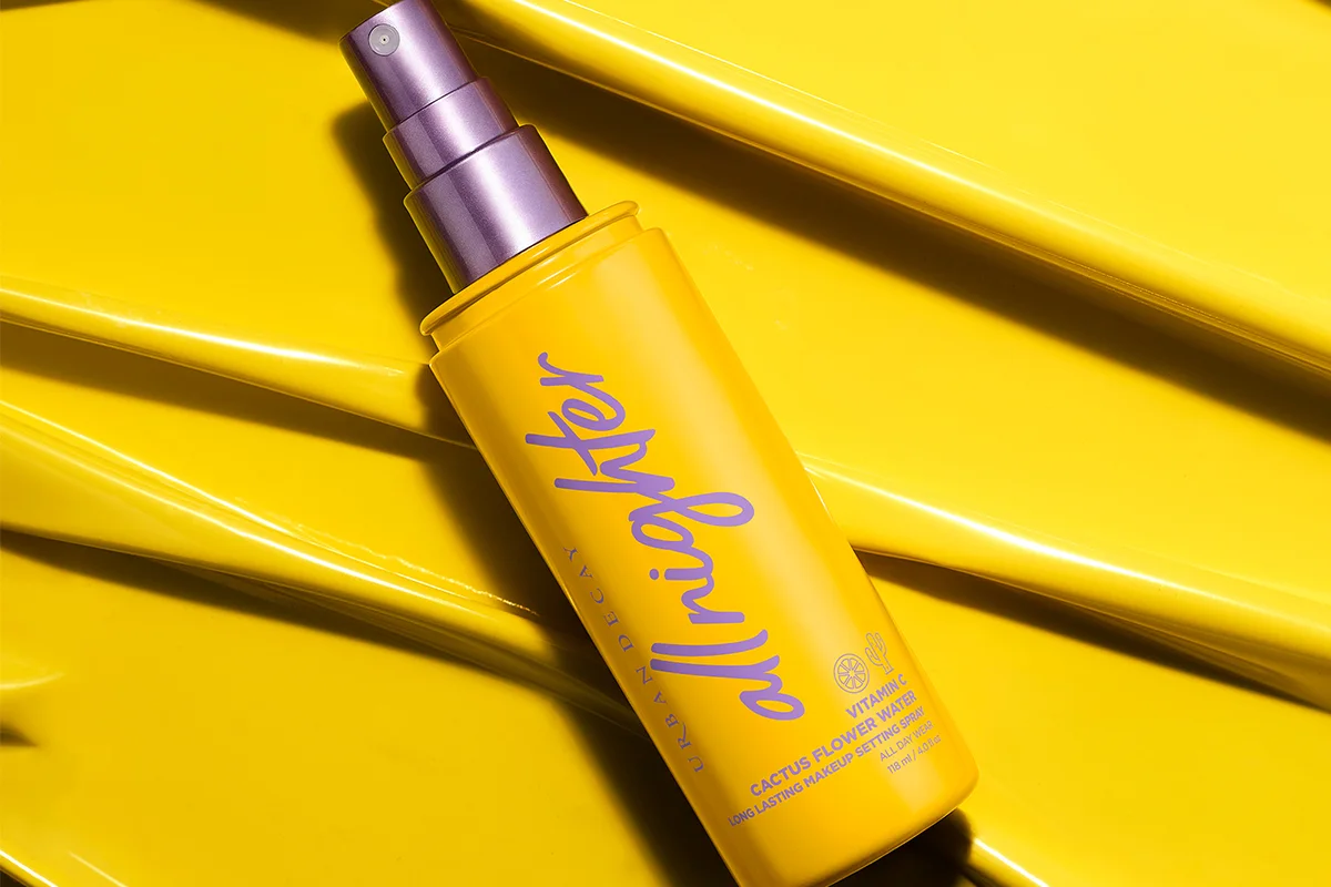 En gul flaske med Urban Decay All Nighter Setting Spray med en lilla spraydyse ligger på en gul flate. Teksten på flasken er lilla.