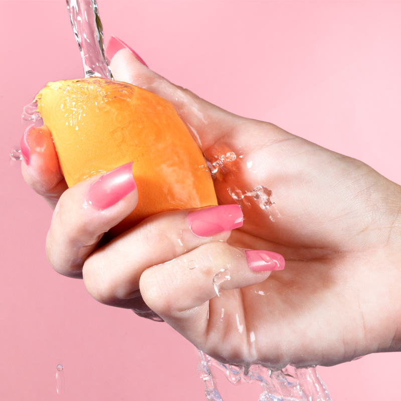 En hånd med rosa neglelakk holder en oransje sminkesvamp under rennende vann mot en rosa bakgrunn. Vannet skaper sprut og drypp rundt hånden og sminkesvampen.