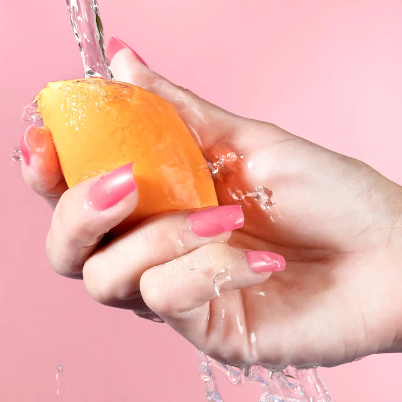 En hånd med rosa neglelakk holder en oransje sminkesvamp under rennende vann mot en rosa bakgrunn. Vannet skaper sprut og drypp rundt hånden og sminkesvampen.