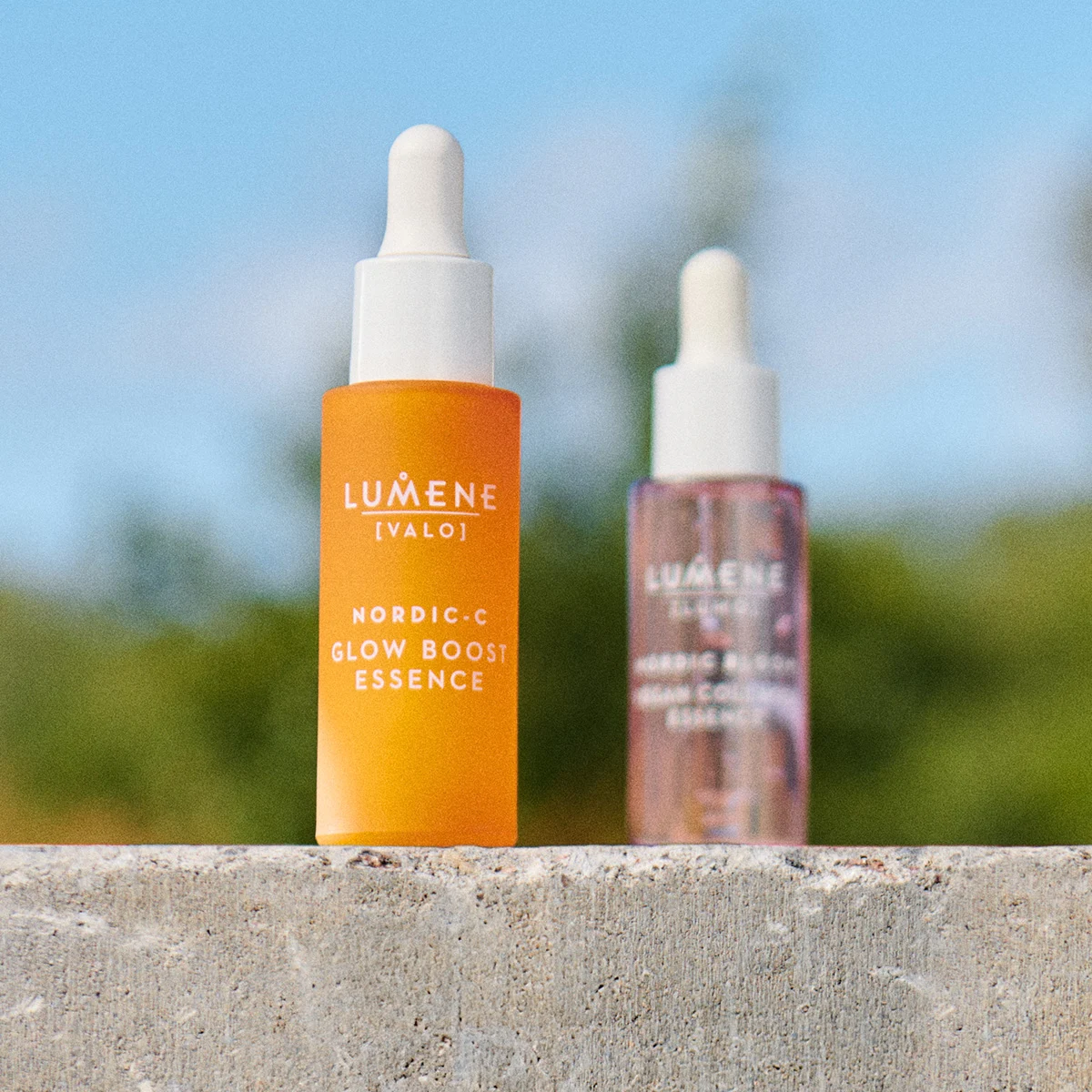 Två flaskor med Lumene hudvårdsprodukter står på en betongyta utomhus. Den främre flaskan är orange och innehåller Nordic-C Glow Boost Essence, medan den bakre flaskan är genomskinlig med rosa vätska.