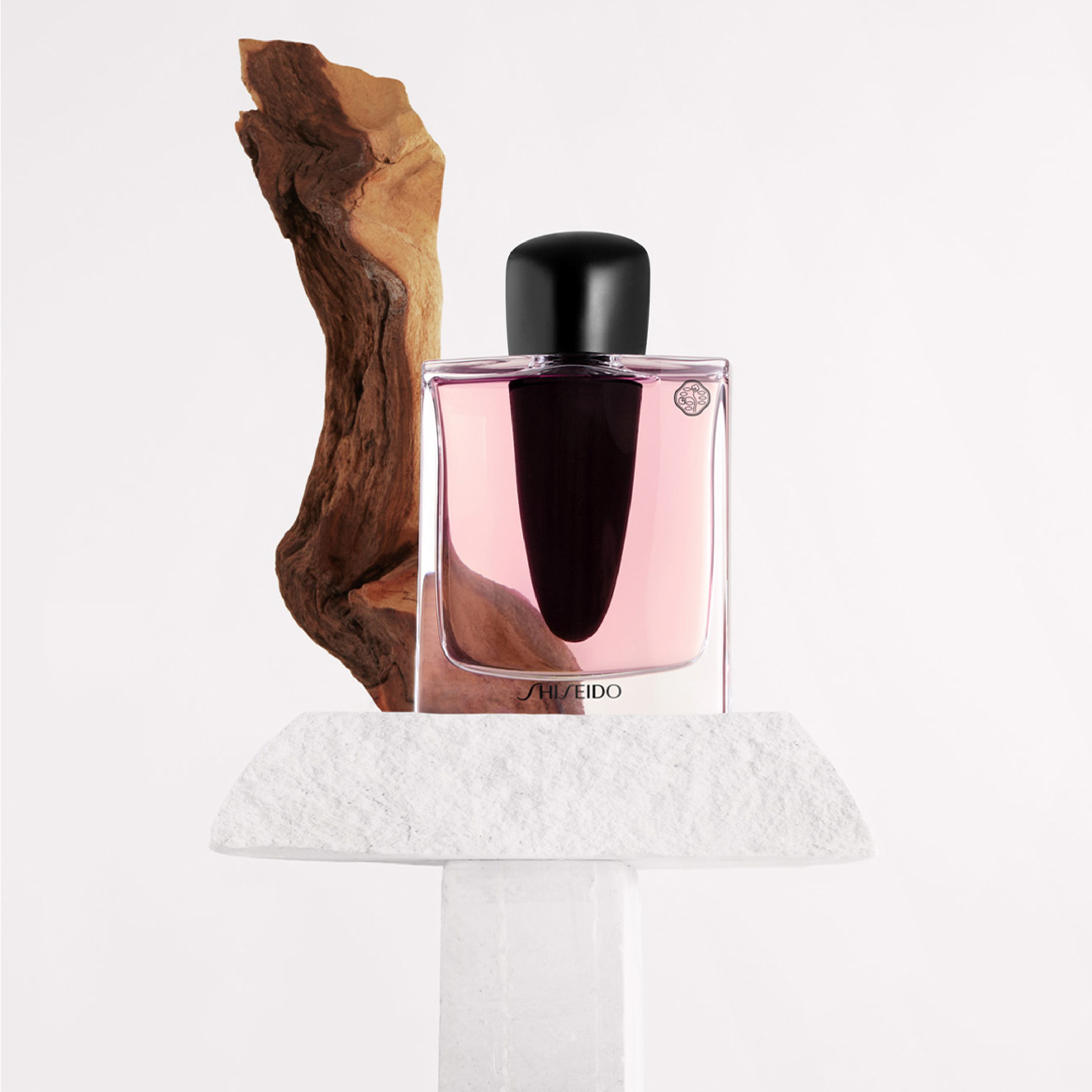 En Shiseido-parfym i en rosa flaska med svart kork, placerad på en vit piedestal med en träbit bredvid.