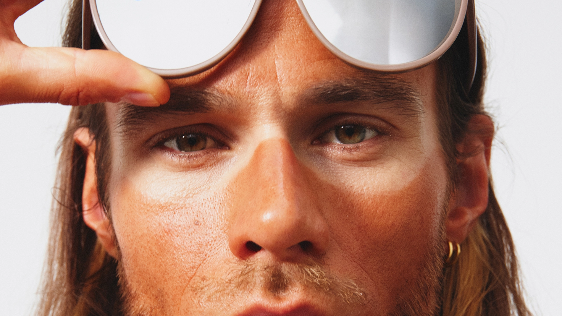 Närbild av ett ansikte där en person lyfter upp ett par solglasögon och pekar på en tydlig ljus rand i pannan efter solglasögonen. Huden visar en ojämn brun-utan-sol-effekt med ljusare område runt ögonen.