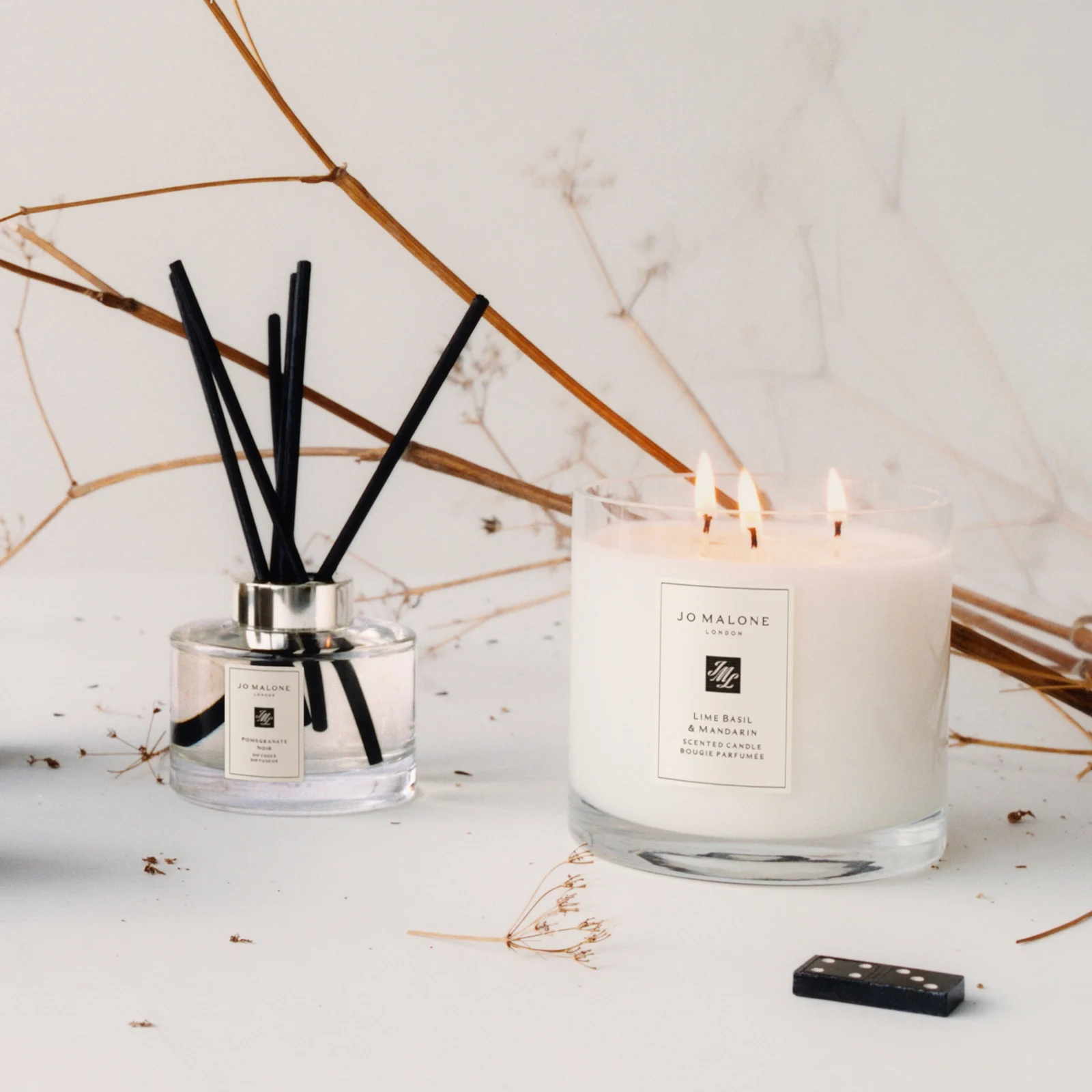 Ett vit doftljus från Jo Malone med texten "Lime Basil & Mandarin" bredvid en doftspridare med svarta pinnar och texten "Pomegranate Noir". Bakgrunden är dekorerad med torra kvistar och blad.