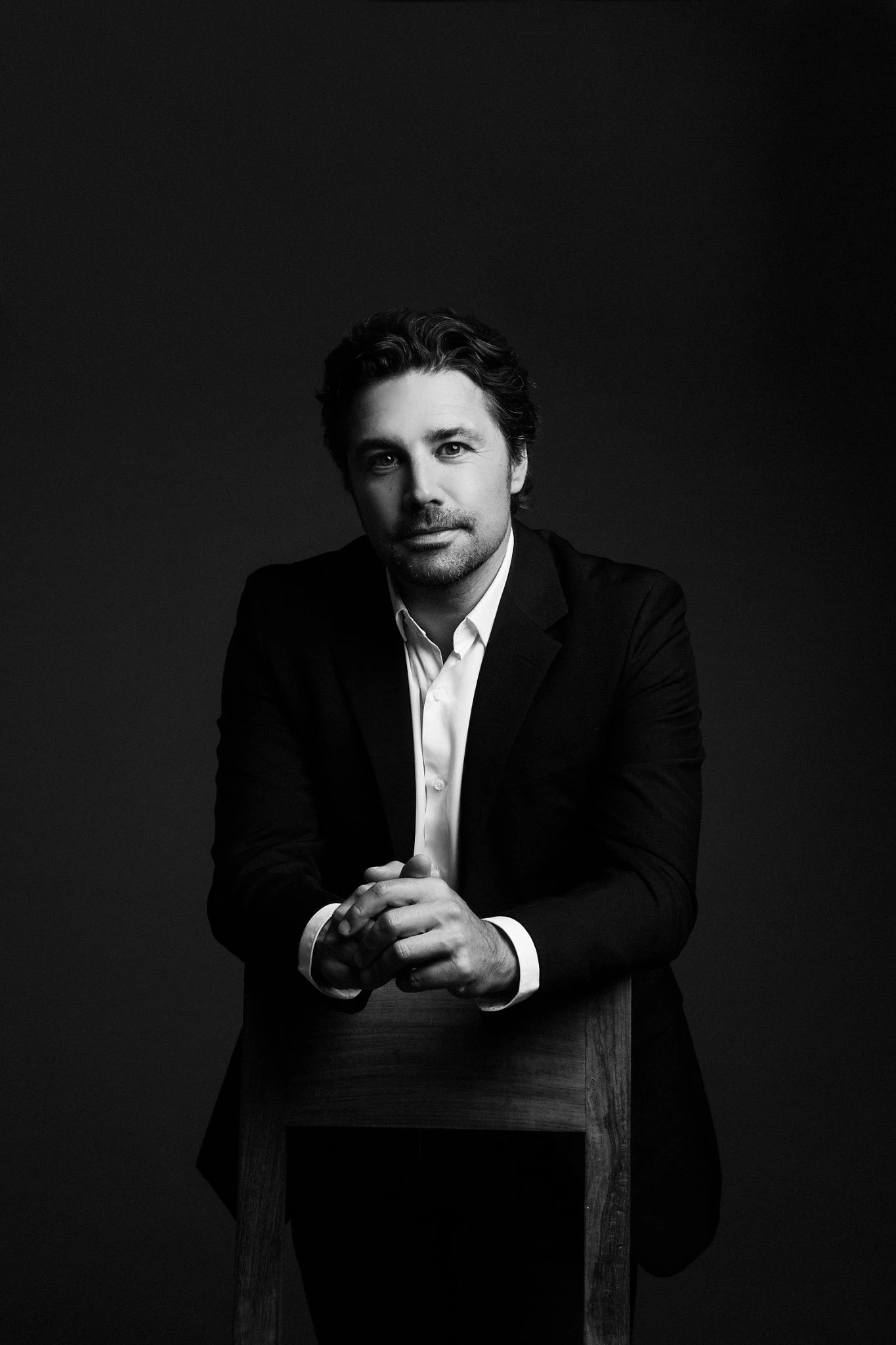 Mustavalkoinen muotokuva henkilöstä, jolla on tumma puku ja vaalea kauluspaita. Henkilö istuu puisen tuolin takana kädet tuolin selkänojan päällä. Tausta on tumma ja yksivärinen pehmeässä valaistuksessa.