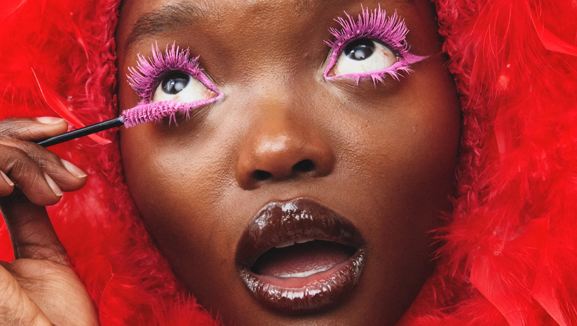 Person påfører intens rosa mascara på de nedre øyevippene med en mascarabørste. Øynene er innrammet med rosa sminke og leppene har mørk, blank lipgloss, mot en rød bakgrunn.