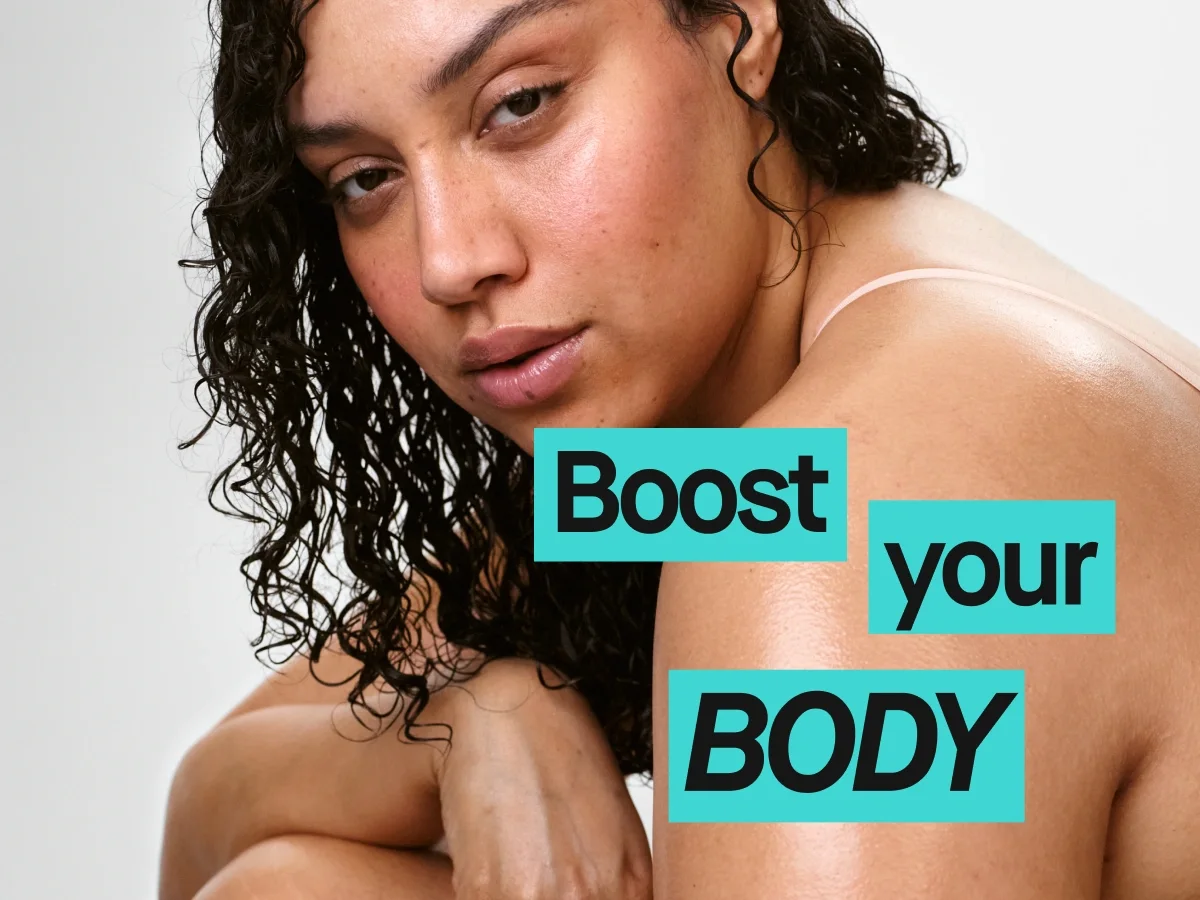 Lähikuva henkilöstä, jolla on tummat kiharat hiukset ja paljas olkapää vaaleaa taustaa vasten. Kuvan päällä näkyy teksti "Boost your BODY", ja painotus on vartalonhoidossa sekä kosteutetun ihon hehkussa.