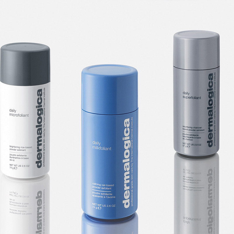 Tre flaskor av exfolieringsprodukter från märket Dermalogica. Från vänster till höger: "Daily Microfoliant" i en vit flaska med grått lock, "Daily Milkfoliant" i en blå flaska, och "Daily Superfoliant" i en grå flaska. Produkterna står på en reflekterande yta som skapar en spegeleffekt. Bakgrunden är ljus och neutral.