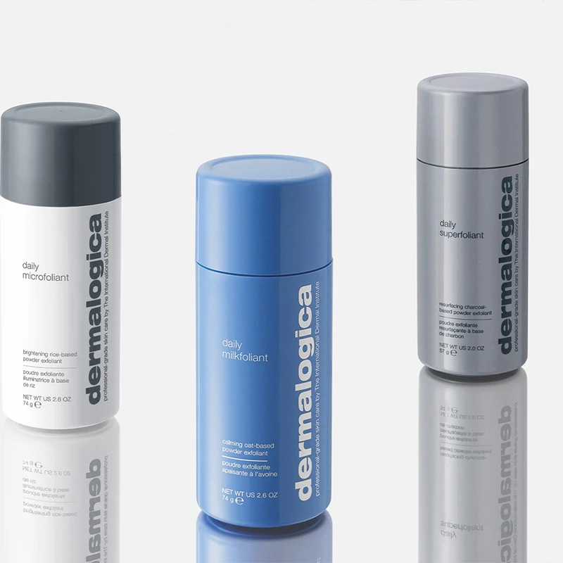 Tre flaskor av exfolieringsprodukter från märket Dermalogica. Från vänster till höger: "Daily Microfoliant" i en vit flaska med grått lock, "Daily Milkfoliant" i en blå flaska, och "Daily Superfoliant" i en grå flaska. Produkterna står på en reflekterande yta som skapar en spegeleffekt. Bakgrunden är ljus och neutral.