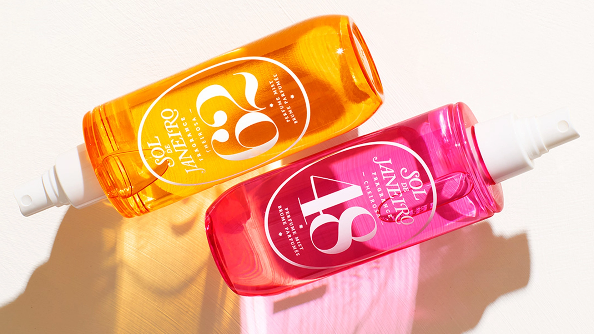 Kaksi värikästä Sol de Janeiro body mist -pulloa makaa valkoisella pinnalla, toinen oranssi ja toinen pinkki.