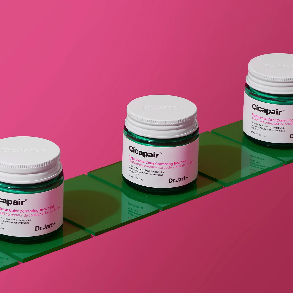 Tre krukker med Dr.Jart+ Cicapair Tiger Grass Colour Correcting Treatment, plassert på grønne fliser mot en rosa bakgrunn.
