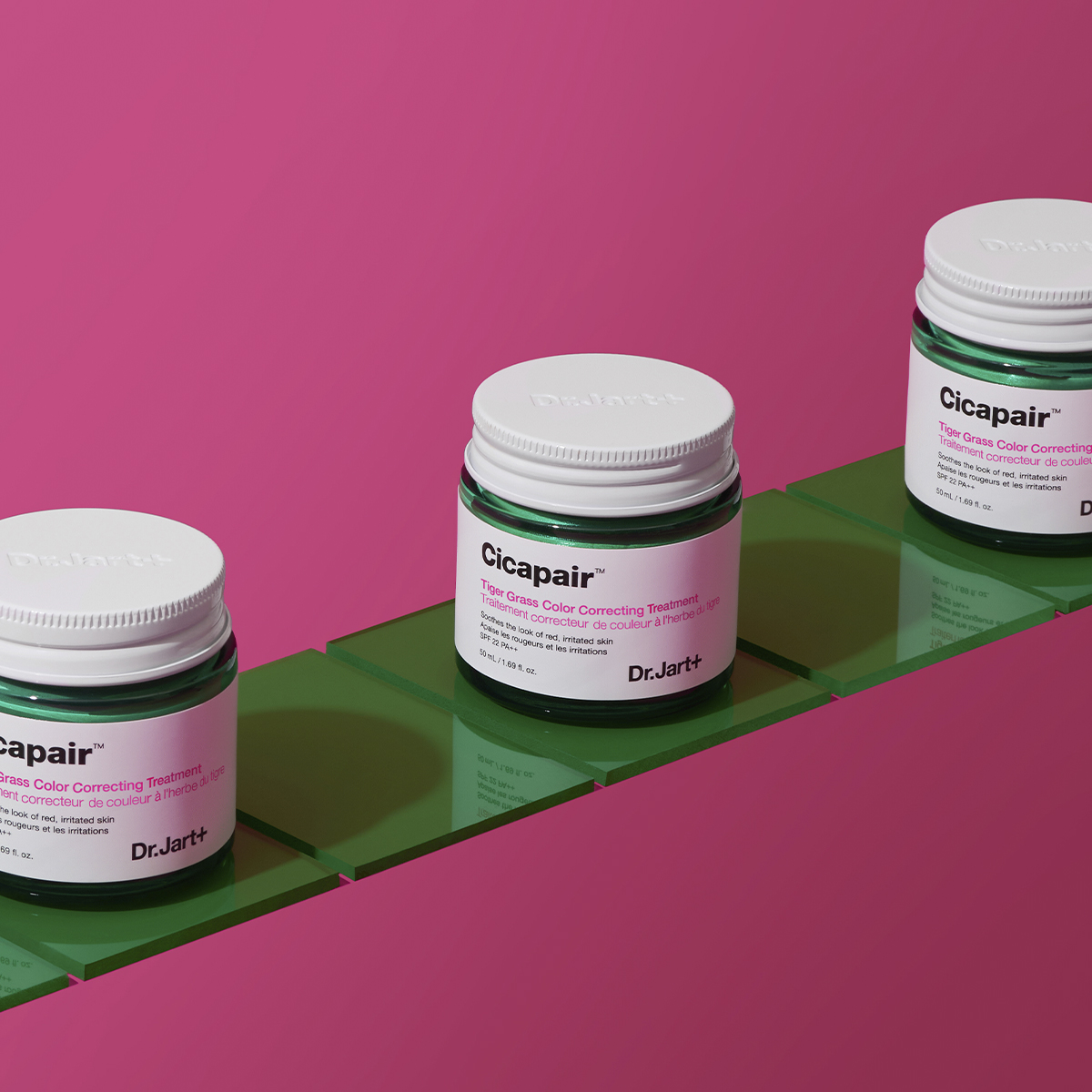 Kolme purkkia Dr.Jart+ Cicapair Tiger Grass Colour Correcting Treatment -värikorjaushoitoa, jotka on sijoitettu vihreille laatoille vaaleanpunaista taustaa vasten.
