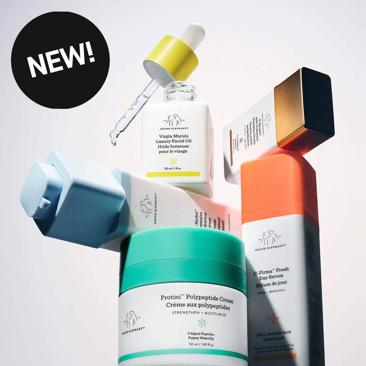 Drunk Elephant hudvårdsprodukter med serum, ansiktsolja och ansiktskräm arrangerade mot ljus bakgrund. Synlig text inkluderar “NEW!”, Virgin Marula Luxury Facial Oil, Protini Polypeptide Cream och C-Firma Fresh Day Serum med färgstarka förpackningar i turkos, orange, gul och blå design.