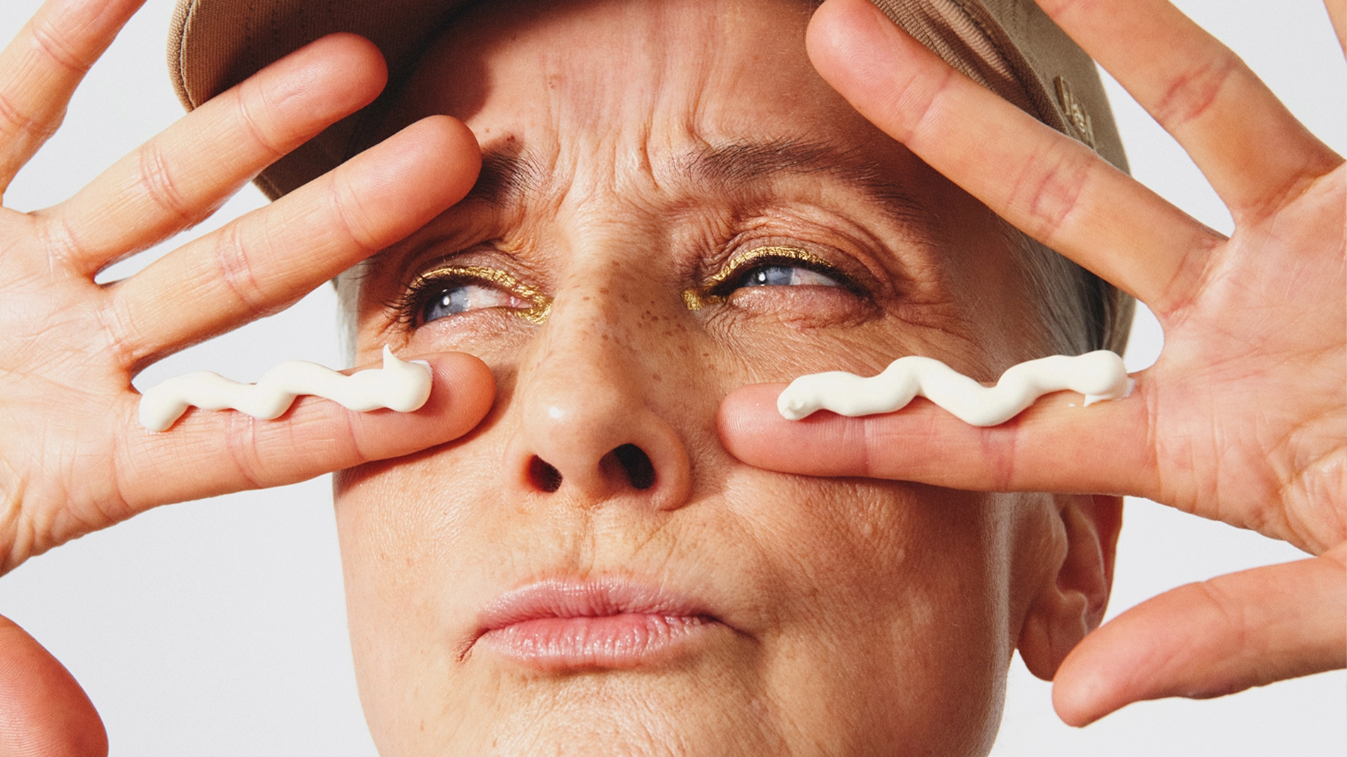 Henkilö, jolla on beige lippalakki, levittää kaksi raitaa silmänympärysvoidetta silmien alle sormenpäillä. Lähikuva kasvoista, kultainen luomiväri ja silmänympärysihon ihonhoito vaaleaa taustaa vasten.