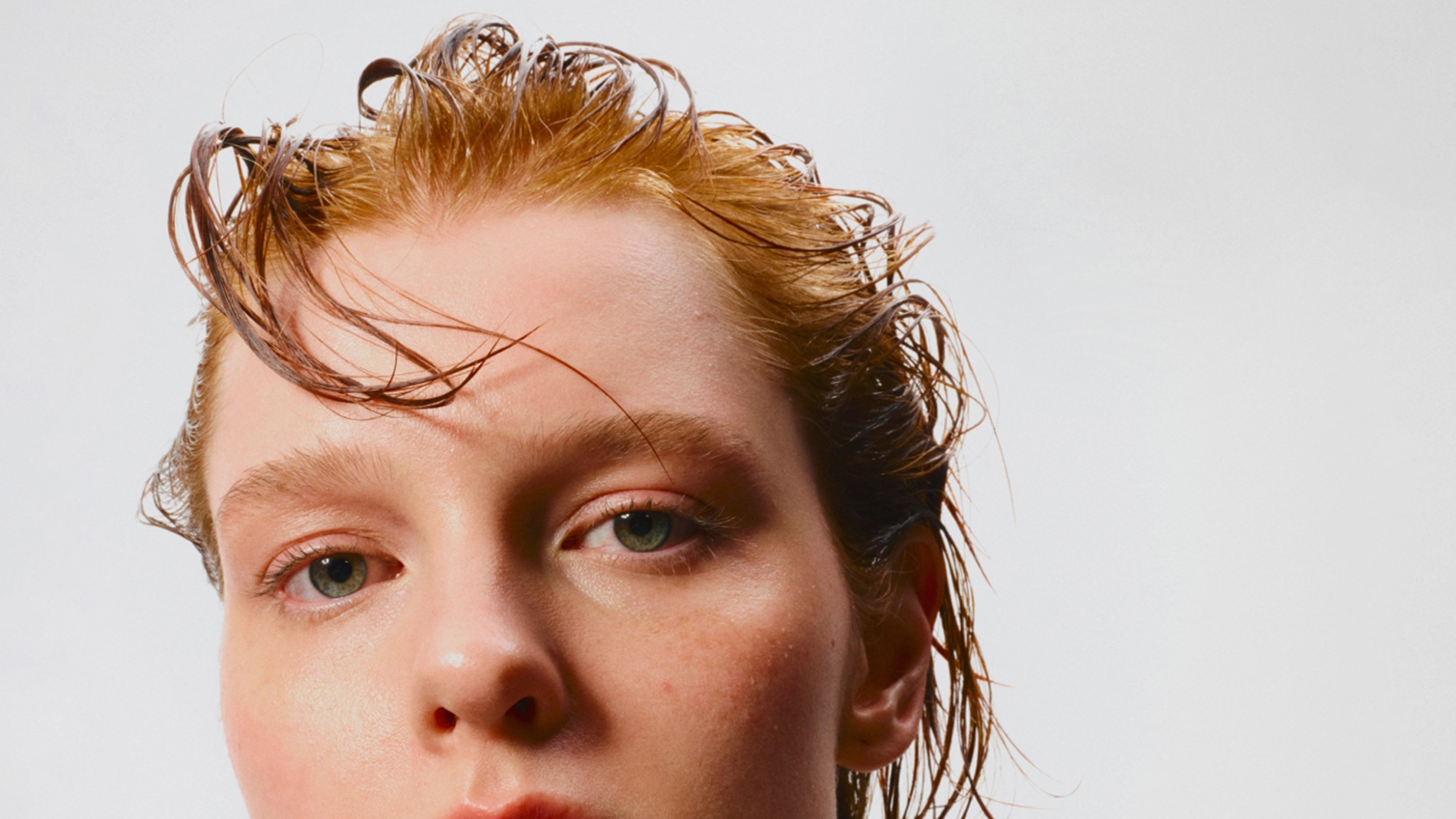 Nærbilde av ansikt med naturlig makeup og fuktig hudfinish, med fokus på hudpleie og basemakeup. Håret er kjemmet bakover og lett vått mot en lys bakgrunn.