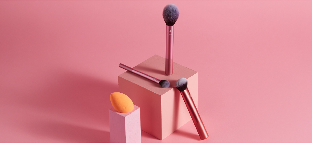 Tre sminkborstar med rosa skaft och en orange makeupsvamp arrangerade på kuber i olika höjder mot en rosa bakgrund, lämpliga för applicering av foundation och puder.