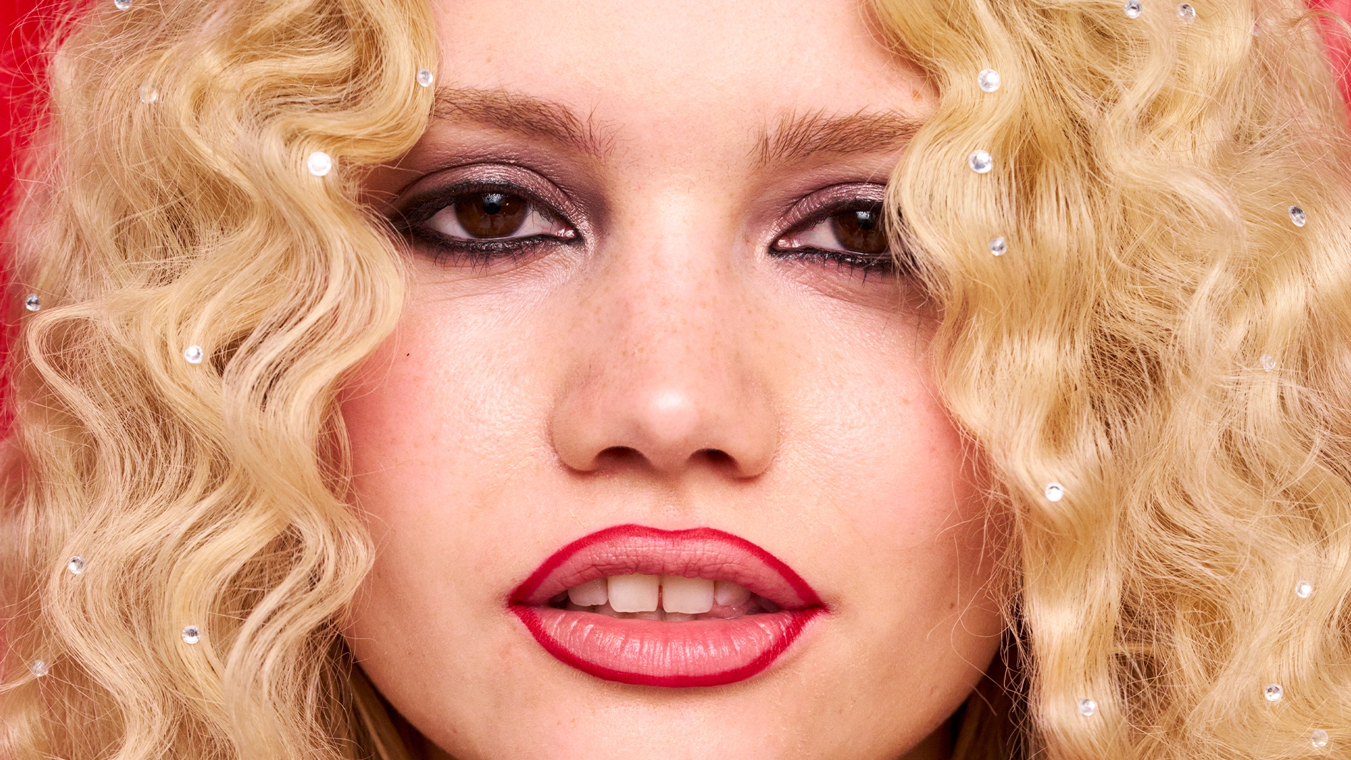 Henkilö, jolla on pitkät kiharat vaaleat hiukset. Huulien ääriviivat on maalattu punaisella huulipunalla. Tausta on punainen. 