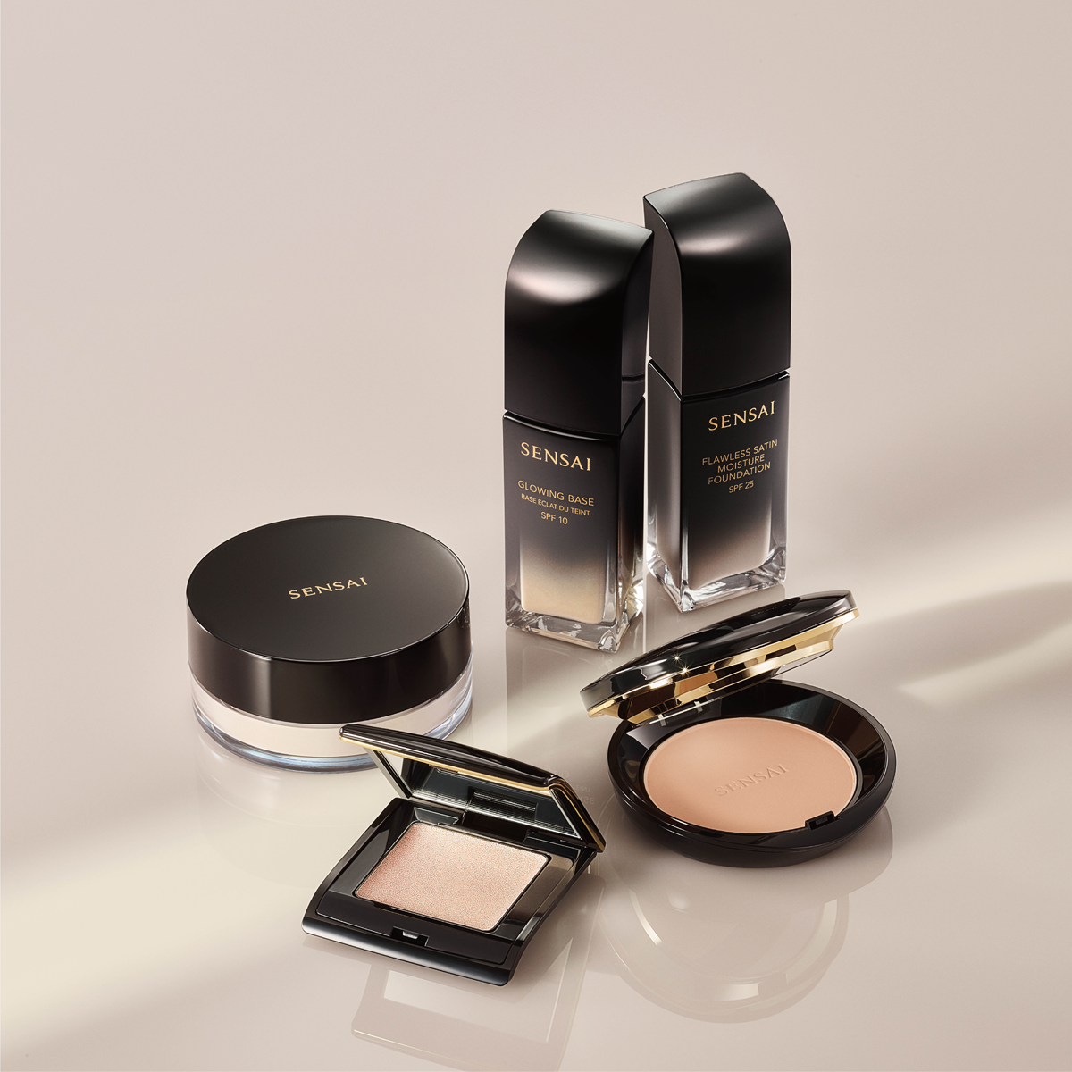 Flere makeup-produkter fra SENSAI vises mot en beige bakgrunn, inkludert Glowing Base SPF 10, Flawless Satin Moisture Foundation SPF 25, pudder, highlighter og compact foundation. Produktene har svart og gullfarget design med reflekterende detaljer og gir et eksklusivt uttrykk av premium foundation og makeup med glow-finish.