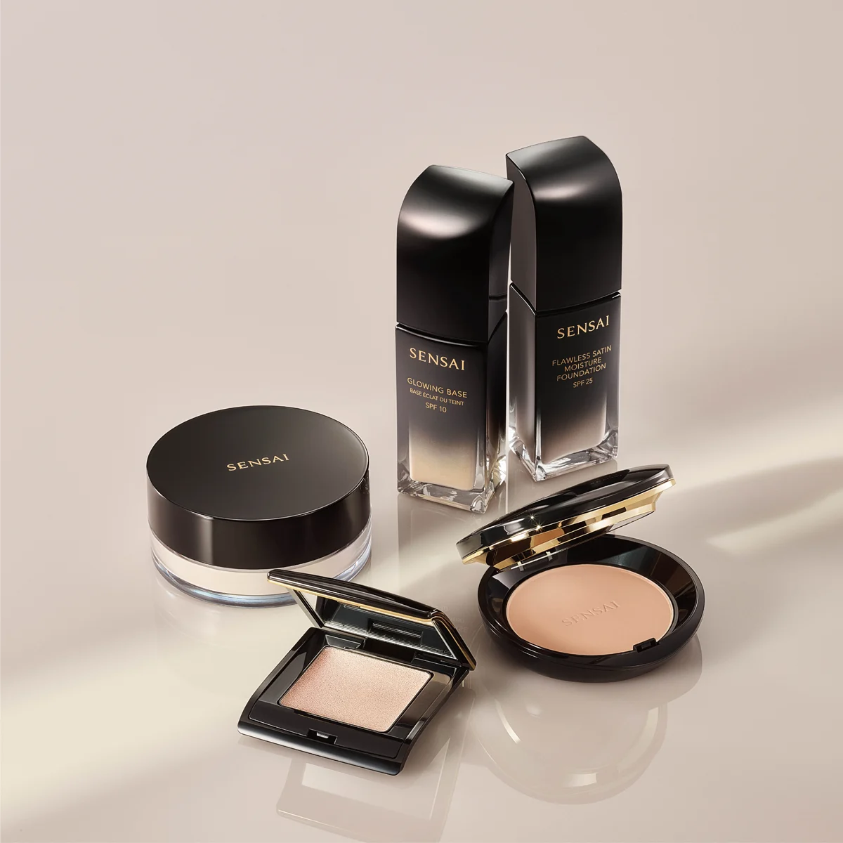 Flera makeup-produkter från SENSAI presenteras mot en beige bakgrund, inklusive Glowing Base SPF 10, Flawless Satin Moisture Foundation SPF 25, puder, highlighter och compact foundation. Produkterna har svart och guldfärgad design med speglande detaljer och ger ett exklusivt intryck av premium foundation och makeup för glow-finish.