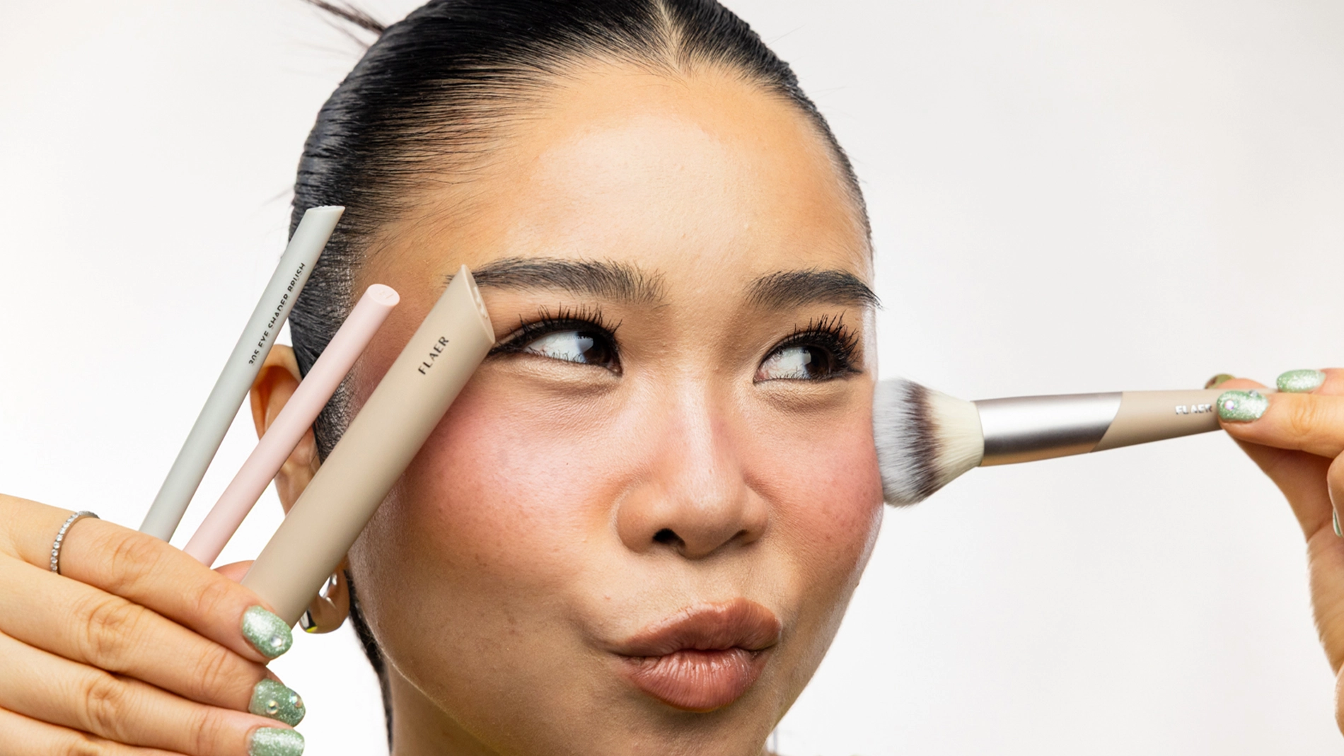 Person med mørkt hår satt opp i lav knute påfører highlighter med sminkekost på kinnbenet. Personen holder flere beige og lyse sminkekoster i én hånd.