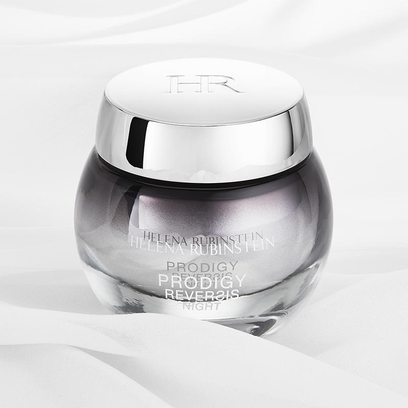 Tyylikäs Helena Rubinstein Prodigy Reversis Night Cream -yövoiteen purkki valkoisella pohjalla, jossa on pehmeät kankaan taitokset. Purkki on pyöreä ja läpinäkyvä, ja siinä on hopeanvärinen korkki, johon on kaiverrettu ”HR”. Voiteen väri on hennon violetin sävyinen, ja purkin tekstissä on tuotteen nimi ja tuotemerkki. Muotoilu on ylellinen ja hienostunut.