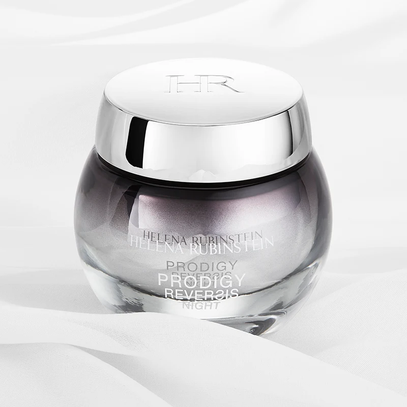 Tyylikäs Helena Rubinstein Prodigy Reversis Night Cream -yövoiteen purkki valkoisella pohjalla, jossa on pehmeät kankaan taitokset. Purkki on pyöreä ja läpinäkyvä, ja siinä on hopeanvärinen korkki, johon on kaiverrettu ”HR”. Voiteen väri on hennon violetin sävyinen, ja purkin tekstissä on tuotteen nimi ja tuotemerkki. Muotoilu on ylellinen ja hienostunut.
