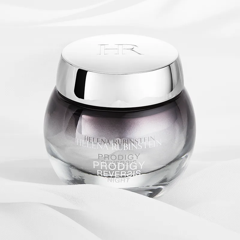 Tyylikäs Helena Rubinstein Prodigy Reversis Night Cream -yövoiteen purkki valkoisella pohjalla, jossa on pehmeät kankaan taitokset. Purkki on pyöreä ja läpinäkyvä, ja siinä on hopeanvärinen korkki, johon on kaiverrettu ”HR”. Voiteen väri on hennon violetin sävyinen, ja purkin tekstissä on tuotteen nimi ja tuotemerkki. Muotoilu on ylellinen ja hienostunut.