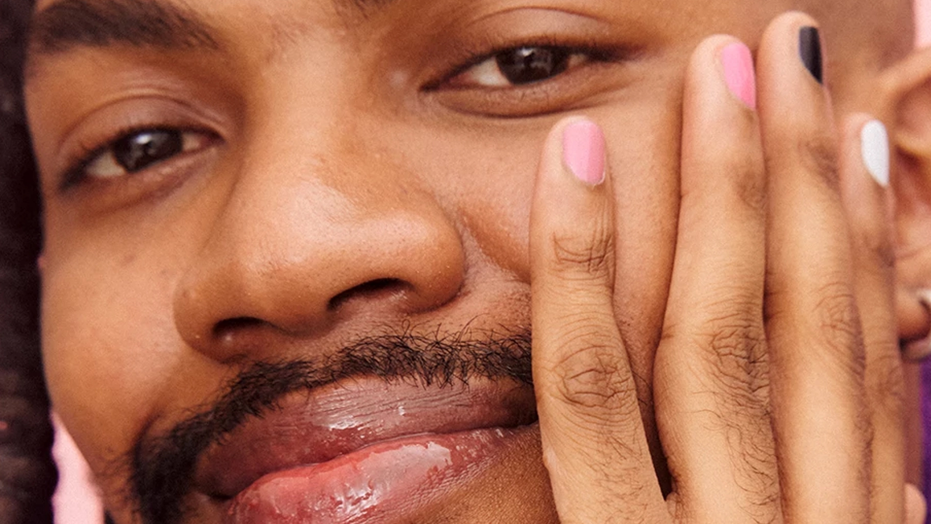 Leende person med färgglatt nagellack och glansiga läppar, poserar mot en rosa bakgrund.