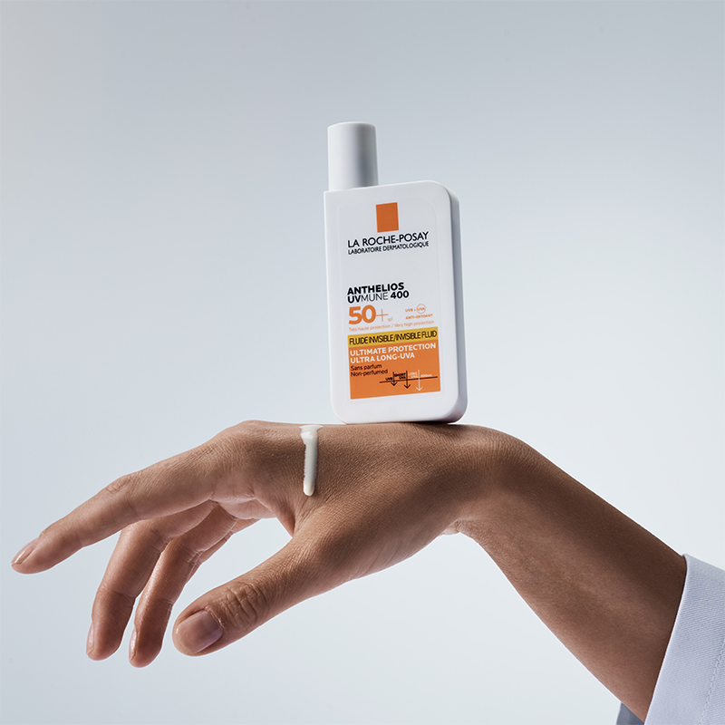En hand håller en flaska La Roche-Posay Anthelios UVmune 400 solskydd med SPF 50+ mot en ljus bakgrund. En liten mängd av solskyddet är applicerad på handens ovansida. Flaskan är vit med orange detaljer och text som beskriver produktens höga solskydd och långvariga UVA-skydd.