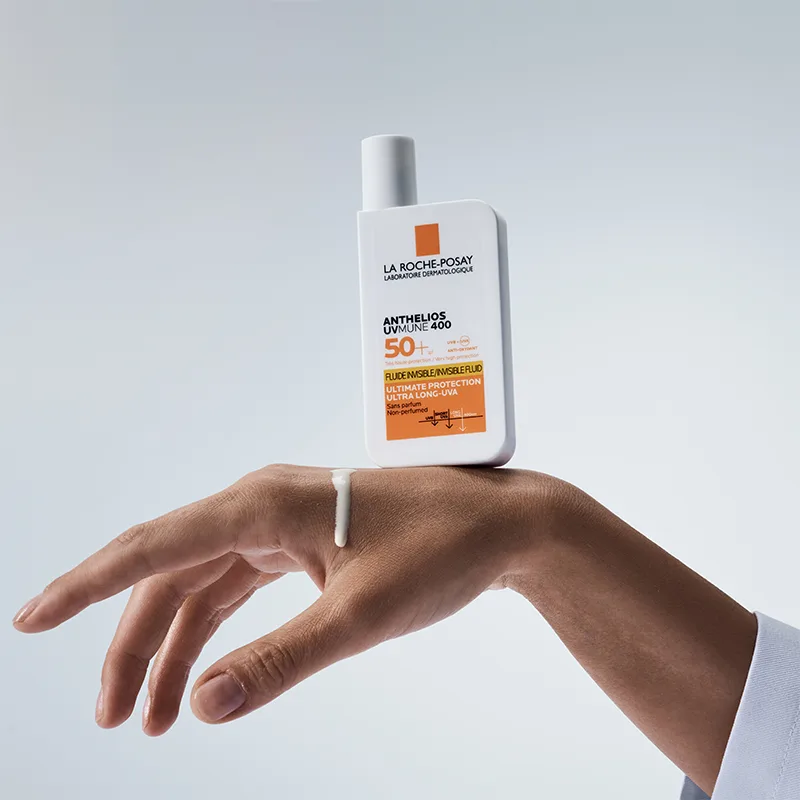 En hand håller en flaska La Roche-Posay Anthelios UVmune 400 solskydd med SPF 50+ mot en ljus bakgrund. En liten mängd av solskyddet är applicerad på handens ovansida. Flaskan är vit med orange detaljer och text som beskriver produktens höga solskydd och långvariga UVA-skydd.