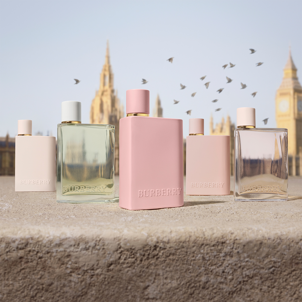 Flere Burberry-parfymeflasker, hvorav en er rosa, plassert på en betongoverflate. I bakgrunnen ses Big Ben og andre bygninger i London, samt flygende fugler.