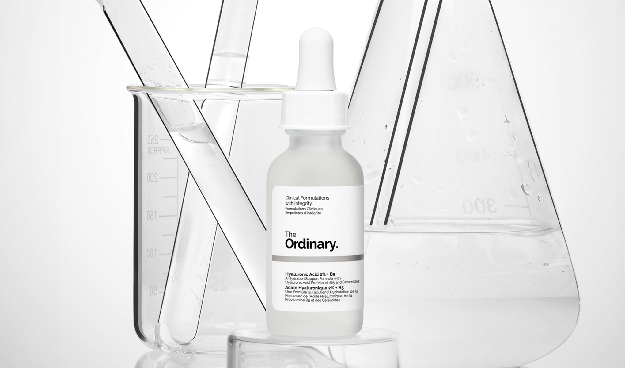 En flaske med pipette plassert foran flere laboratorieglass med klar væske. Bakgrunnen er hvit og ren, noe som understreker produktets vitenskapelige og kliniske preg. Emballasjen er enkel og stilren med en hvit etikett og svart tekst.