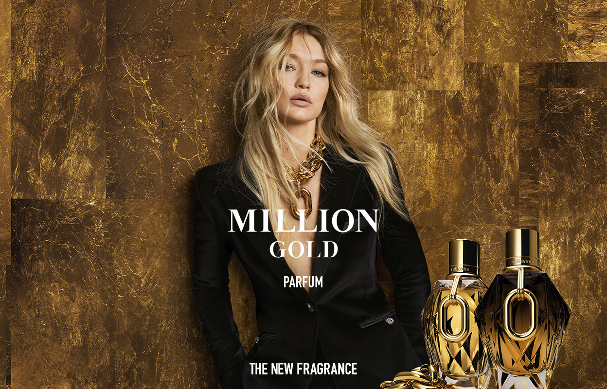 En kvinna med långt blont hår står framför en gyllene bakgrund. Hon bär en svart kavaj och ett stort halsband. Framför henne står två flaskor av parfymen "Million Gold". Texten på bilden lyder: "Million Gold Parfum. The New Fragrance".