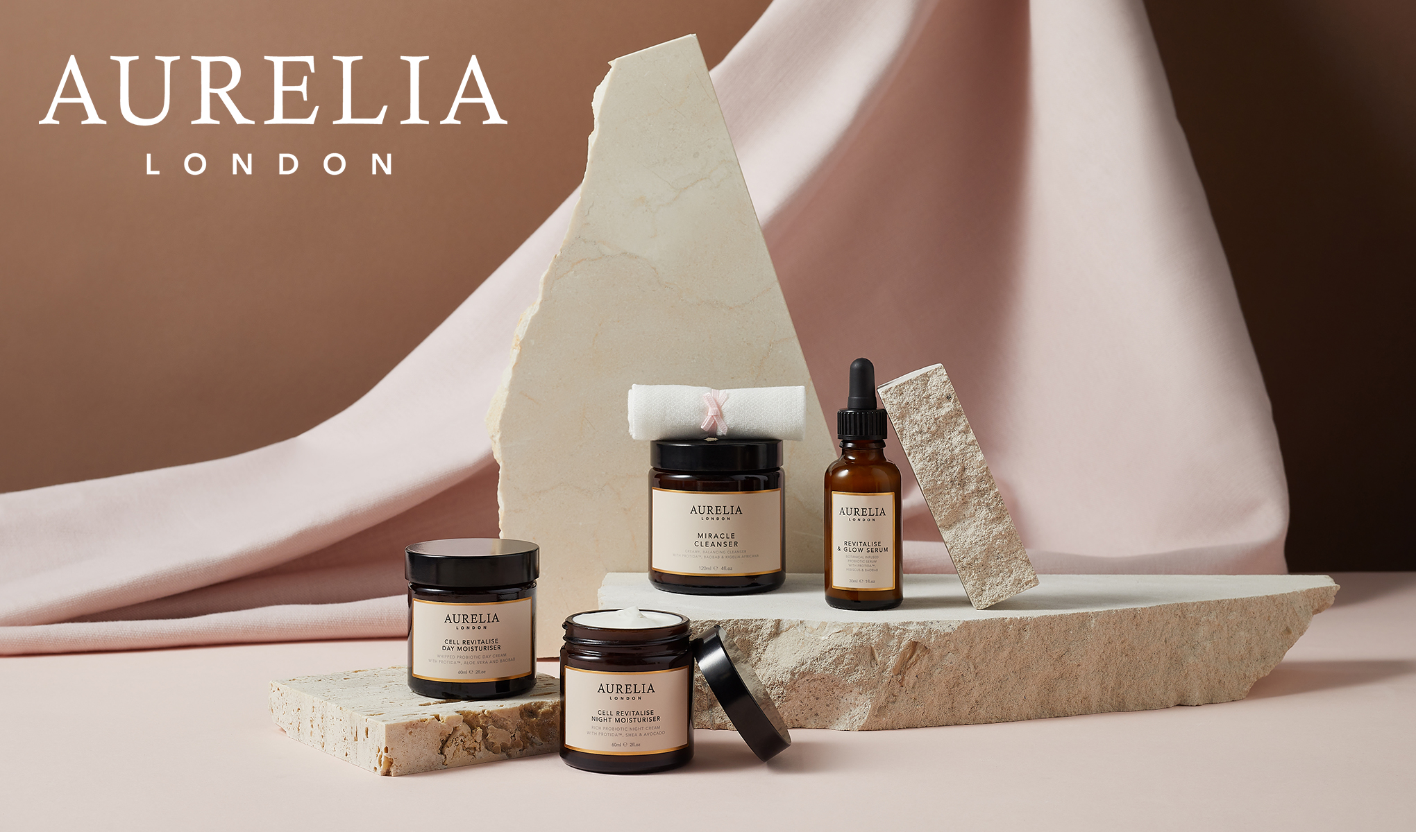 Aurelia London -merkin ihonhoitotuotteiden kokoelma. Tuotteet on aseteltu tyylikkäästi kivien ja vaaleanpunaisen kangastaustan päälle. Tuotemerkin nimi, Aurelia London, näkyy vasemmassa yläkulmassa.