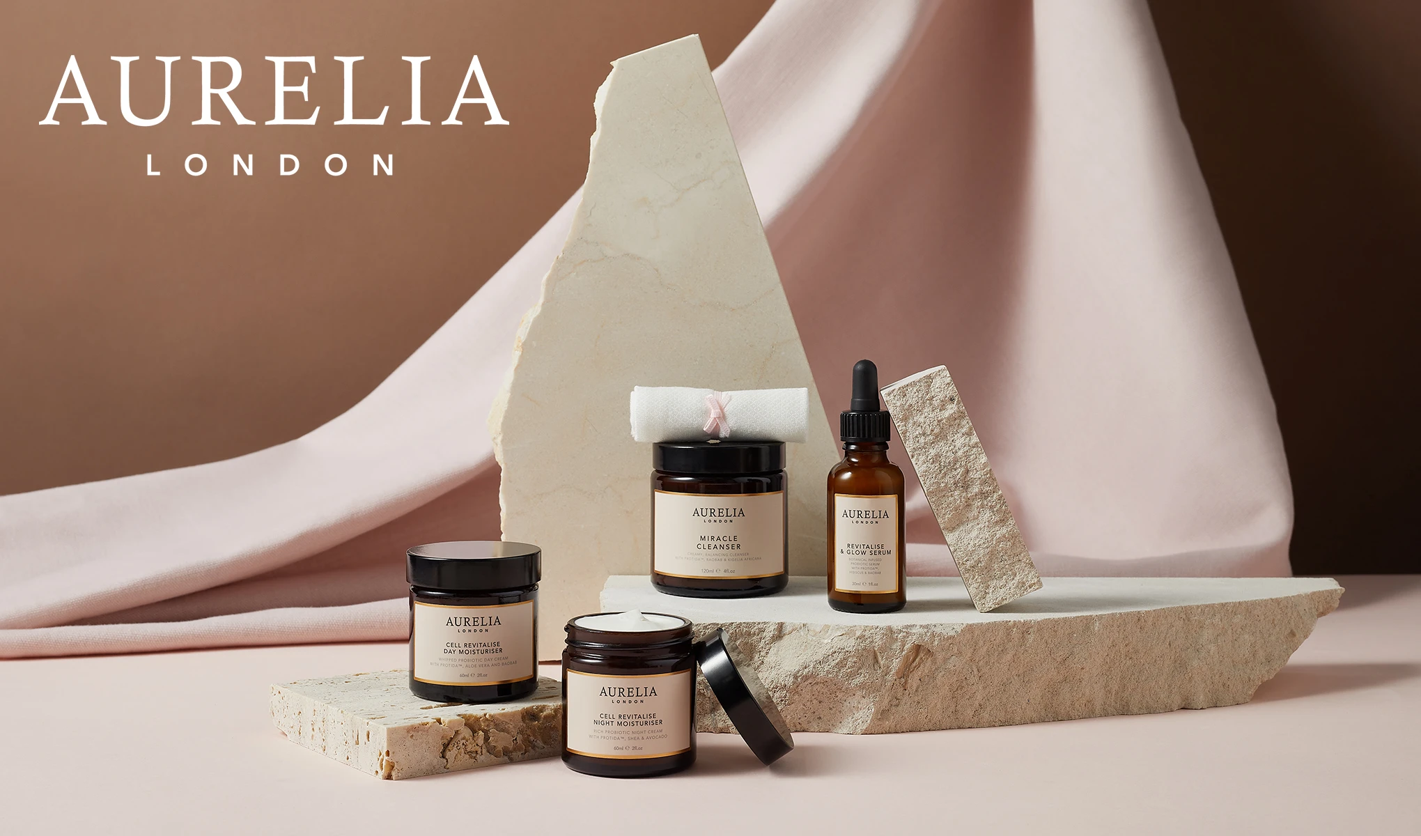 Hudpleieprodukter fra merket Aurelia London. Produktene er elegant arrangert på steiner og en lys rosa stoffbakgrunn. Merkets navn, Aurelia London, er synlig i øvre venstre hjørne.