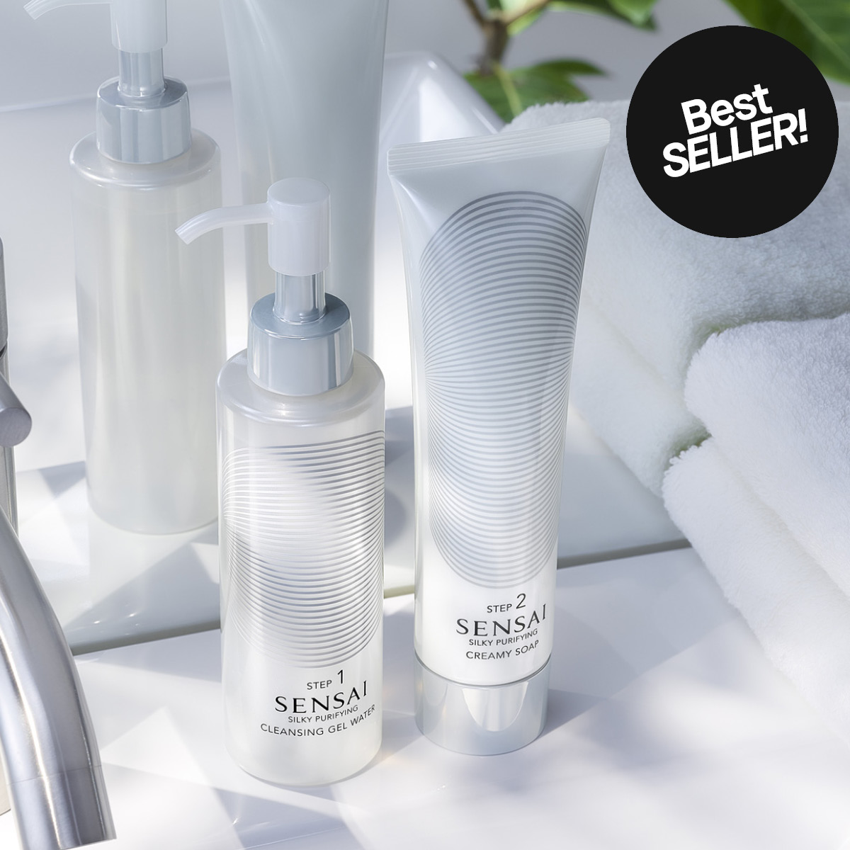 Hudpleieprodukter fra Sensai Silky Purifying står på en servant ved siden av håndklær og plante. Tekst på emballasjen viser “STEP 1 SENSAI CLEANSING GEL WATER” og “STEP 2 SENSAI SILKY PURIFYING CREAMY SOAP” samt en sirkel med “Best SELLER!”.