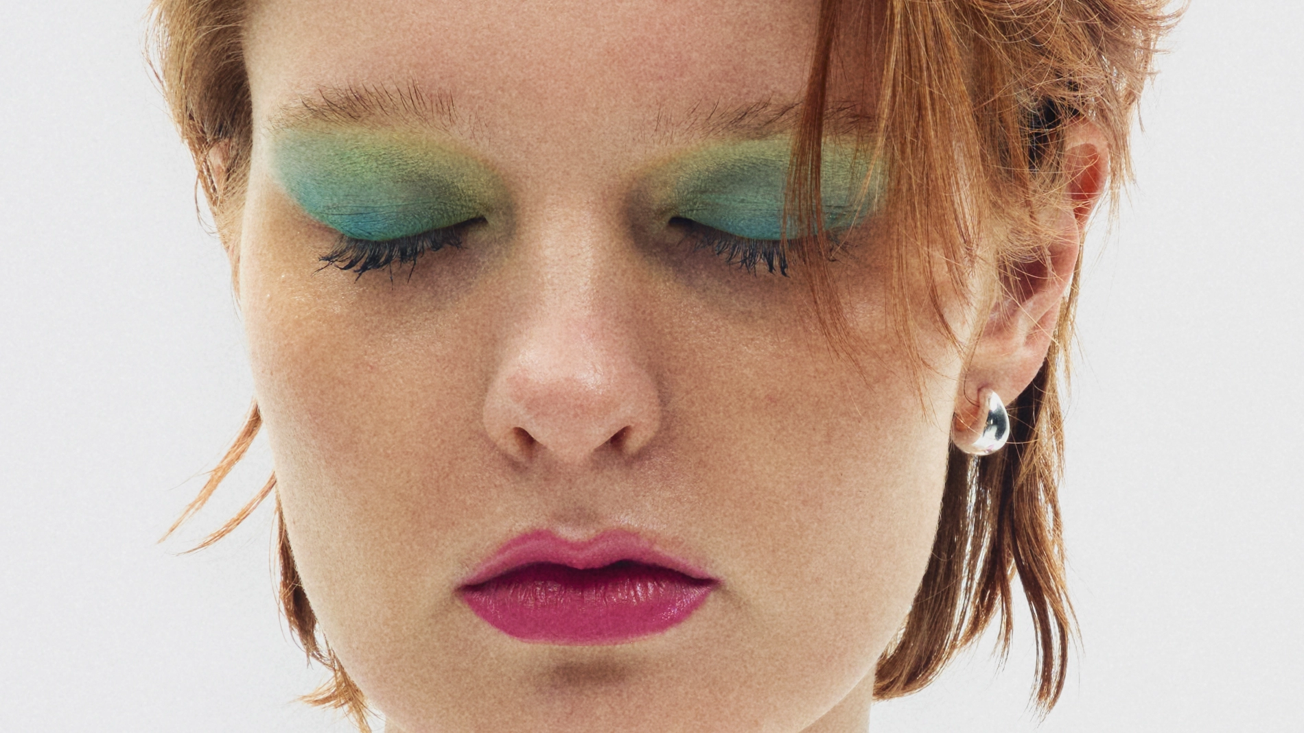 Närbild på person med stark ögonskugga i grönt och blått samt rosa läppstift, ögonen slutna.