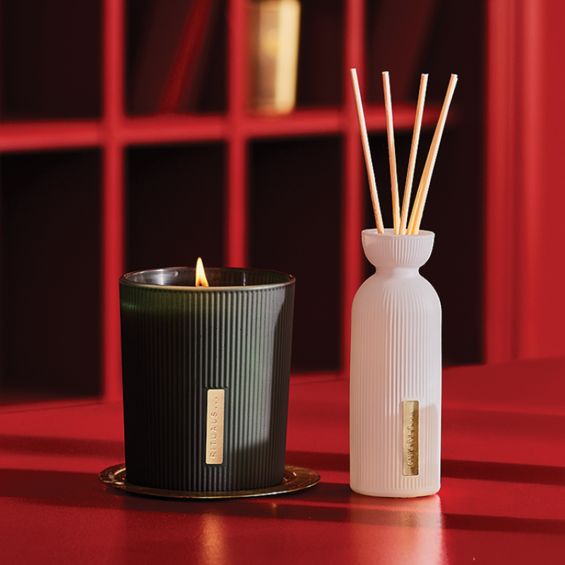 Musta Rituals-kynttilä ja valkoinen tuoksutikkuastia asetettu punaisen taustan eteen. Kynttilä palaa ja tuoksutikkuastia sisältää useita puisia tikkuja.