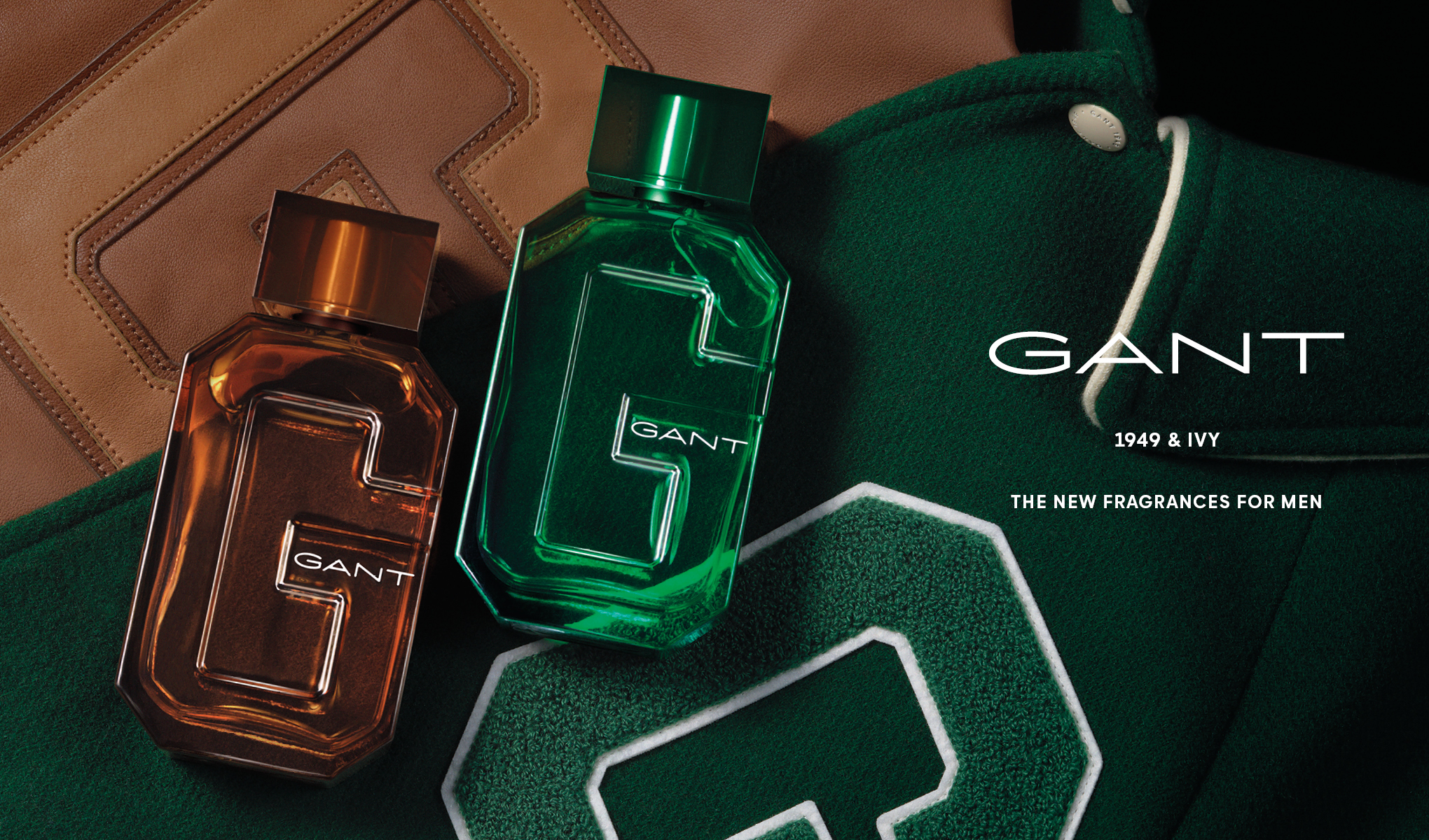 Kaksi GANTin hajuvesipulloa, toinen ruskea ja toinen vihreä, on aseteltu vihreälle kangastaustalle, jossa on nahkaisia yksityiskohtia. Teksti ”GANT 1949 & Ivy The New Fragrances For Men” on sijoitettu pullojen oikealle puolelle. Pulloissa on tyylikäs muotoilu ja niihin on kaiverrettu suuri G-kirjain. Tausta ja pullot luovat tyylikkään ja hienostuneen esityksen.