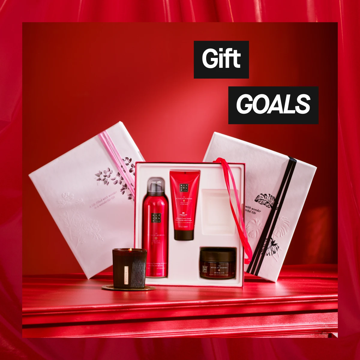 Rituals-lahjapakkaus valkoisissa laatikoissa, punainen sisäosa ja satiininauha, sisältää punaisia vartalotuotteita ja palavan kynttilän, aseteltuna punaiselle pöydälle punaista taustaa vasten – tyylikäs joululahjakuva.