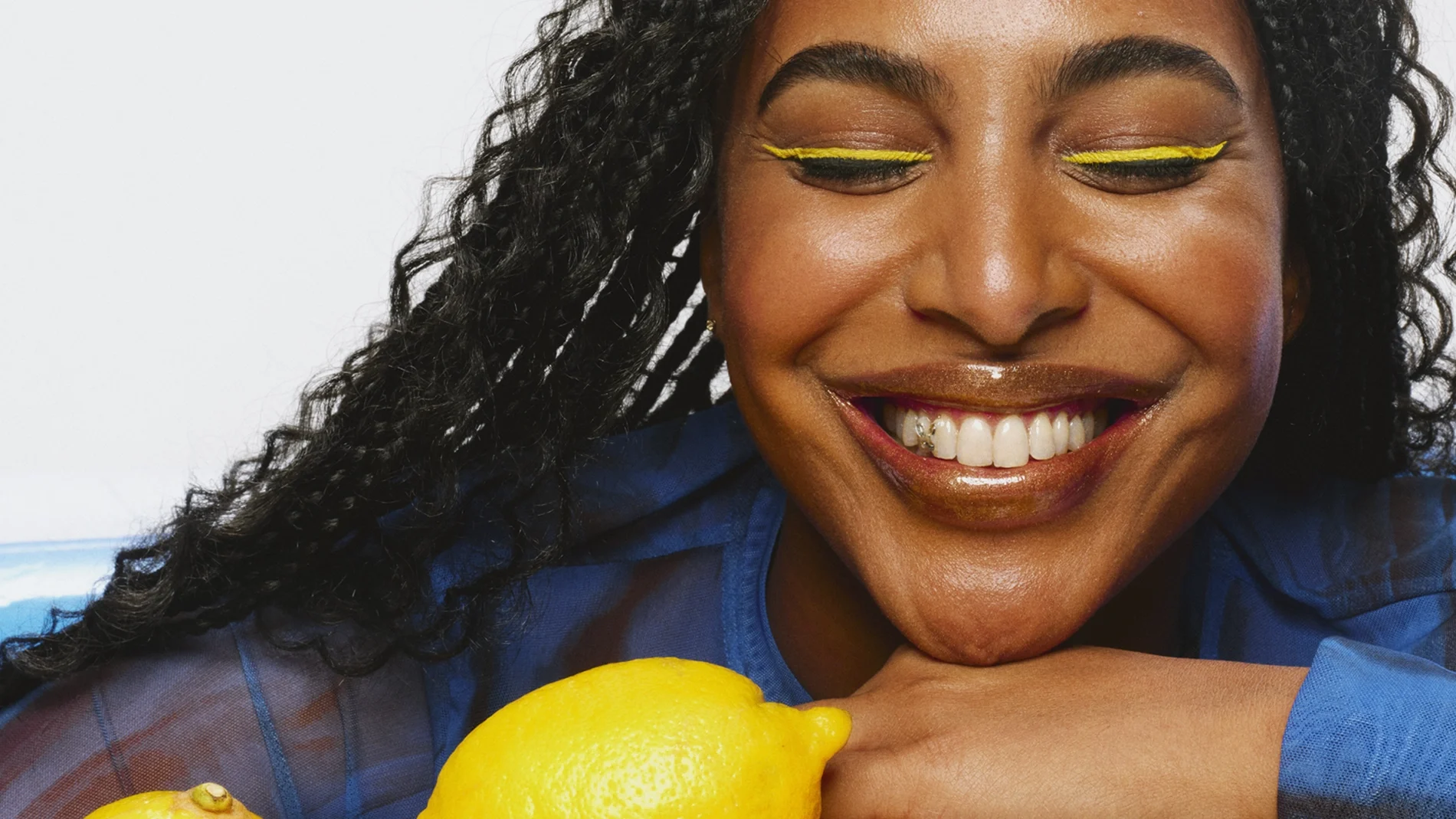 Leende person med flätat hår och färgglad makeup, inklusive gul eyeliner och glansiga bruna läppar. Håller armarna över en hög av citroner.