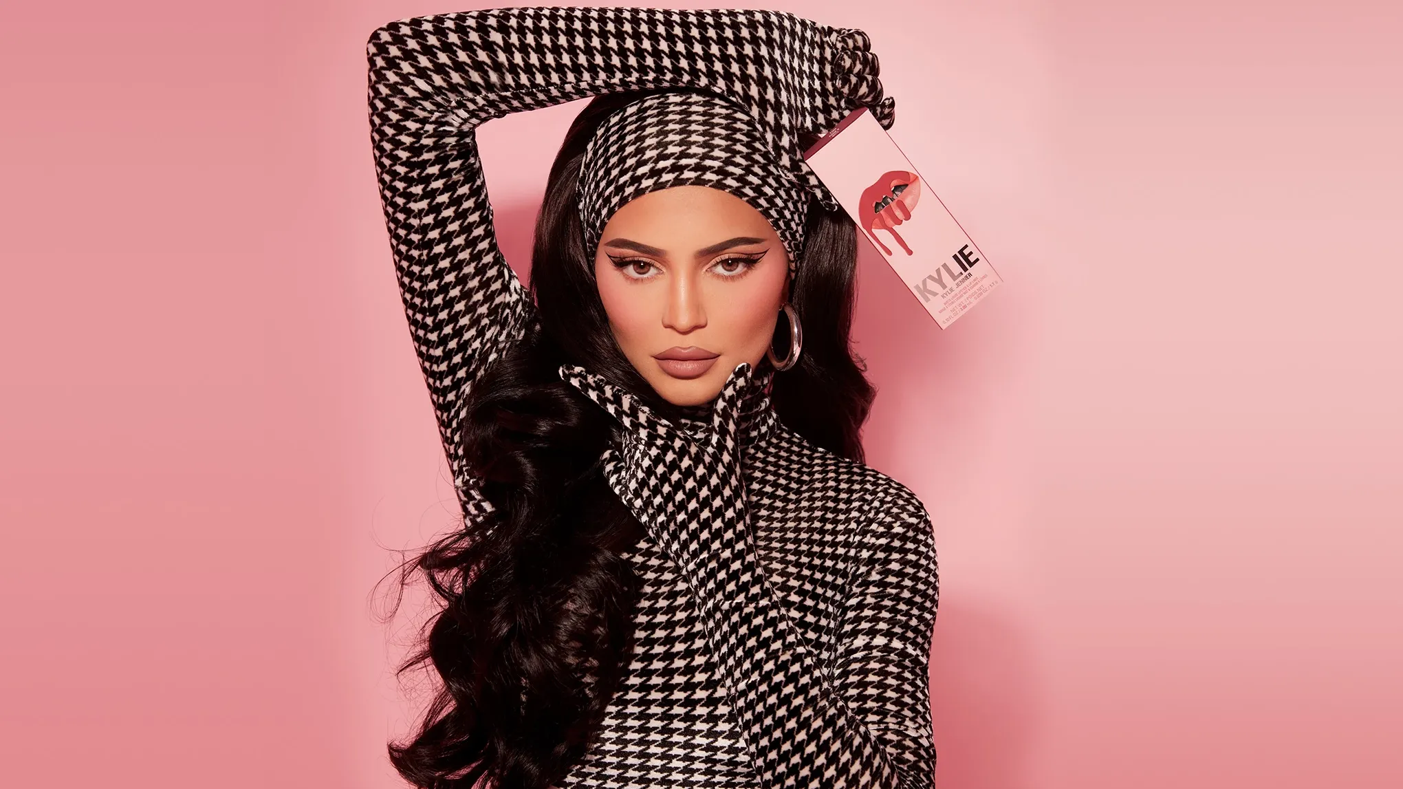 Kylie Jenner poserer foran en rosa bakgrunn i svart-hvitt hundetannsmønstret antrekk og holder opp en eske fra sitt merke, Kylie Cosmetics.