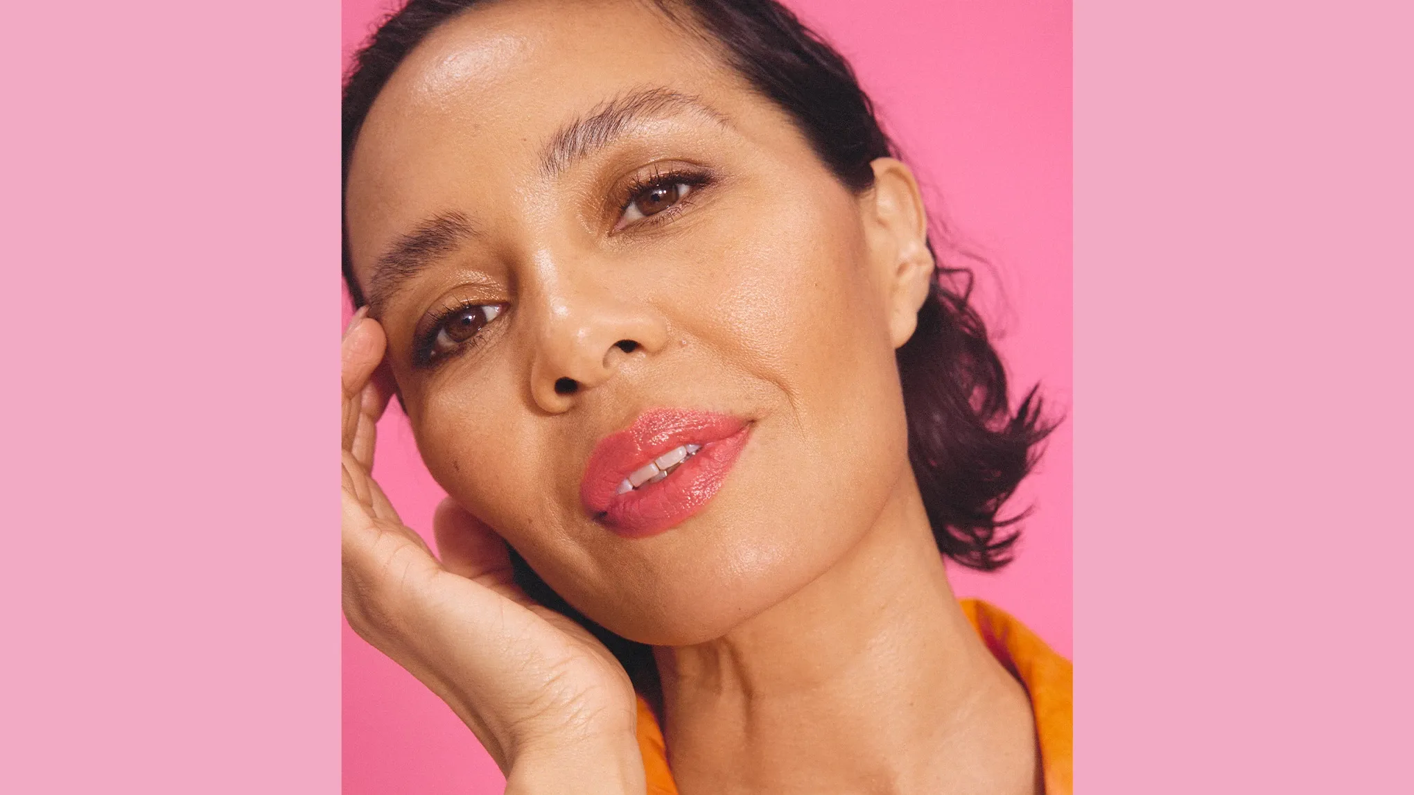 Leende kvinna med kort, svart hår, smink och läppstift, mot en rosa bakgrund.
