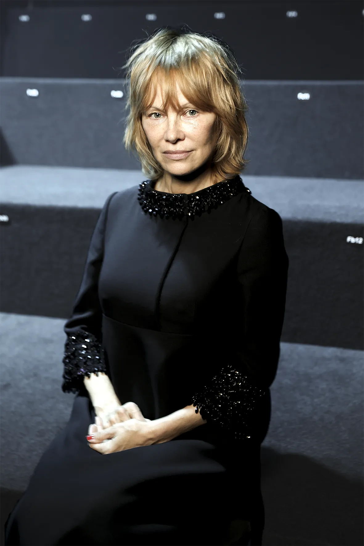 Portrett av en person med kort, lysebrunt hår og naturlig makeup som sitter på grå trappetrinn, iført en svart kjole med dekorative detaljer ved krage og ermer. Personen holder hendene i fanget og ser rett mot kamera i et stilrent og dempet miljø.