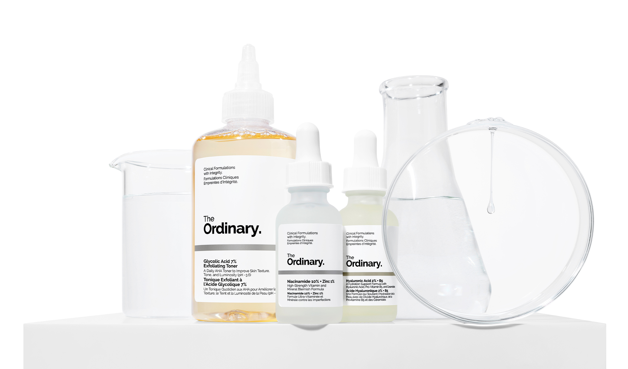 The Ordinary -tuotteita, mukaan lukien kuoriva kasvovesi, niasiiniamidiseerumi ja hyaluronihapposeerumi, aseteltuna valkoiselle taustalle laboratoriovälineiden kanssa.
