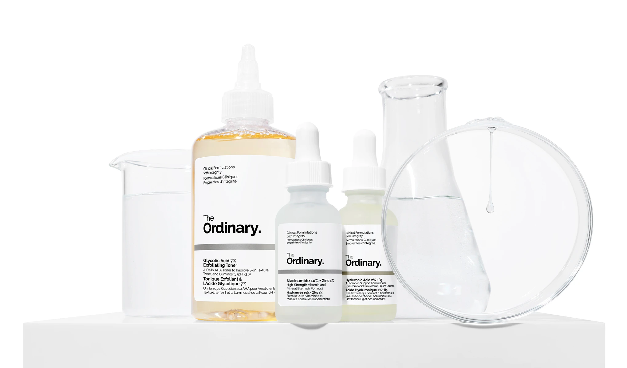 The Ordinary -tuotteita, mukaan lukien kuoriva kasvovesi, niasiiniamidiseerumi ja hyaluronihapposeerumi, aseteltuna valkoiselle taustalle laboratoriovälineiden kanssa.