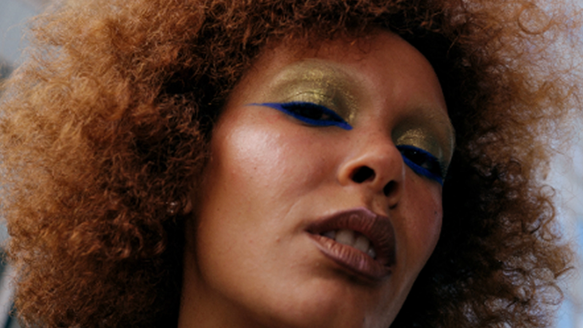 Närbild av person med volymrikt lockigt hår och makeup med guldfärgad ögonskugga och tydlig blå eyeliner, samt naturlig bas och nude läppar.
